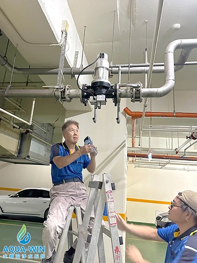 Professional technician on a ladder installing a high-flow automatic backwashing water filter onto a large DN80 overhead pipe in an industrial parking facility.